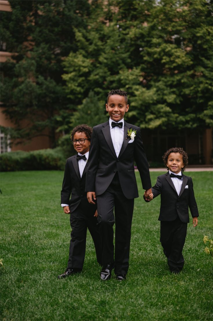 Three young boys in black tuxedos walking together on a green lawn at a wedding