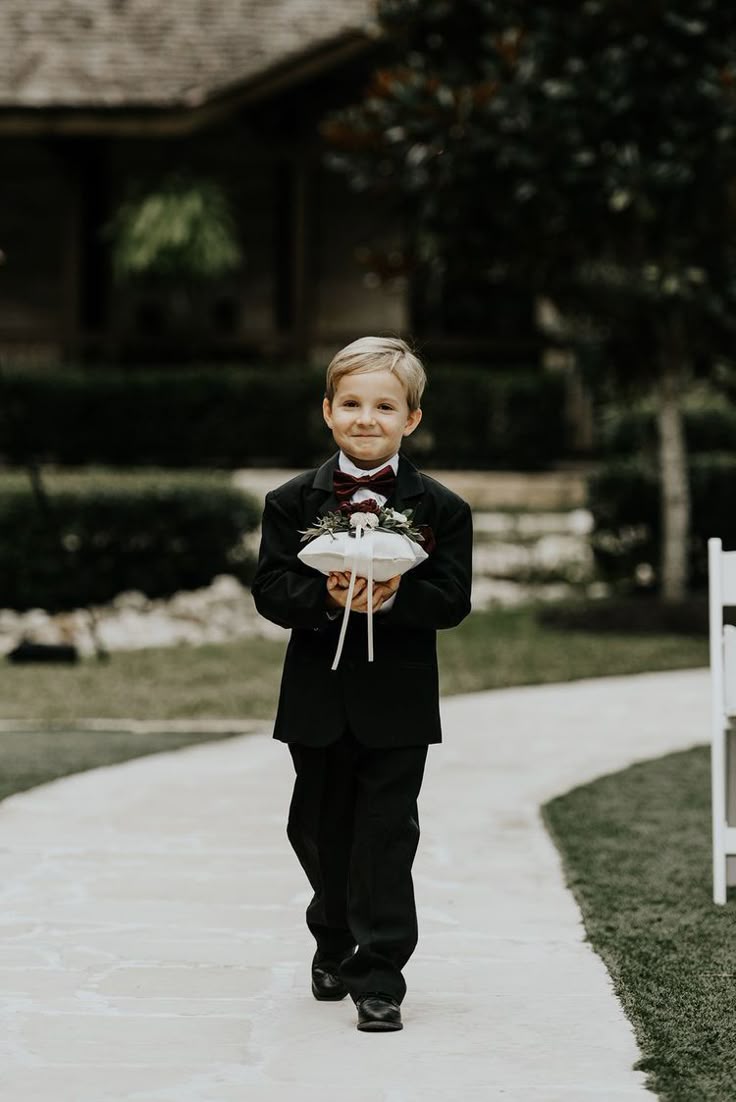 A young ring bearer in a black tuxedo carrying the ring pillow down the aisle